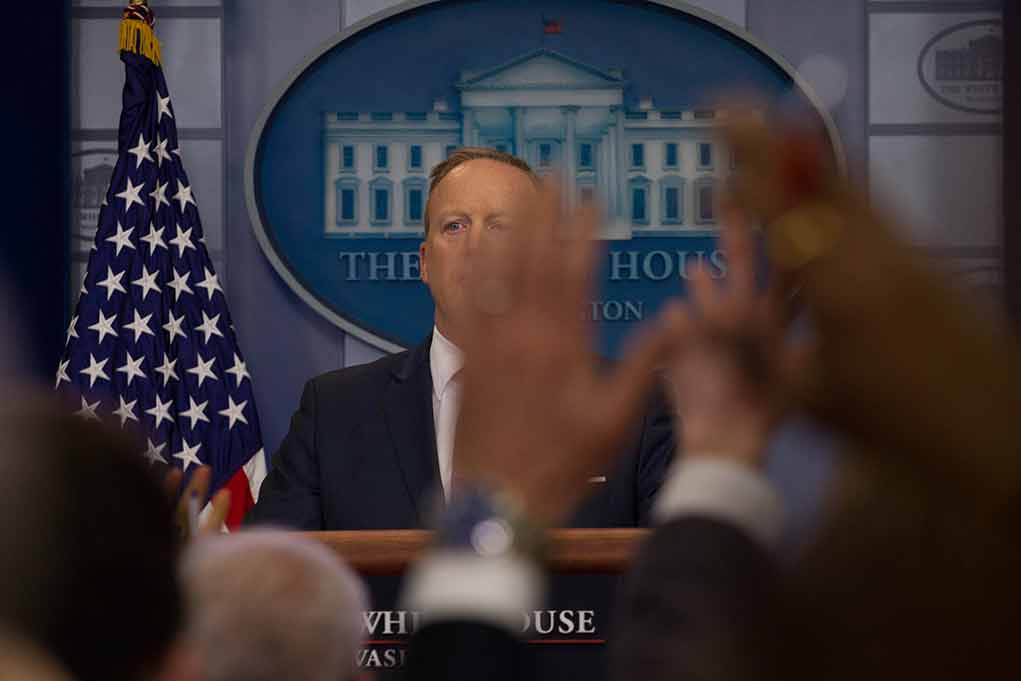 Press secretary at podium with raised hands in briefing room